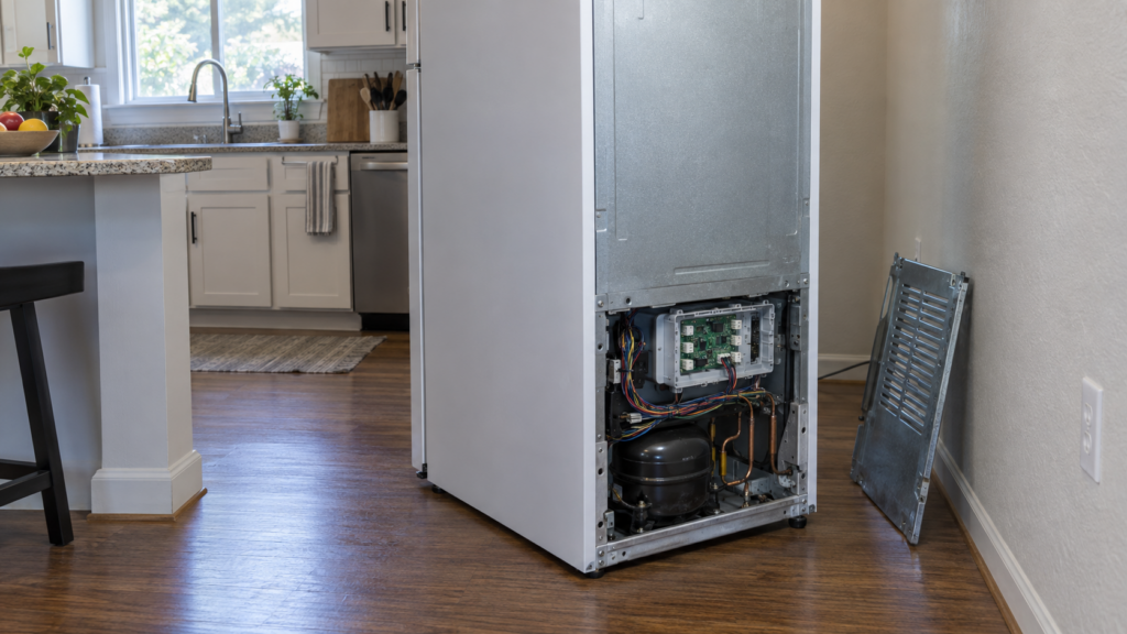 A standalone upright freezer pulled from the wall, its rear access panel removed to reveal the control board housing.